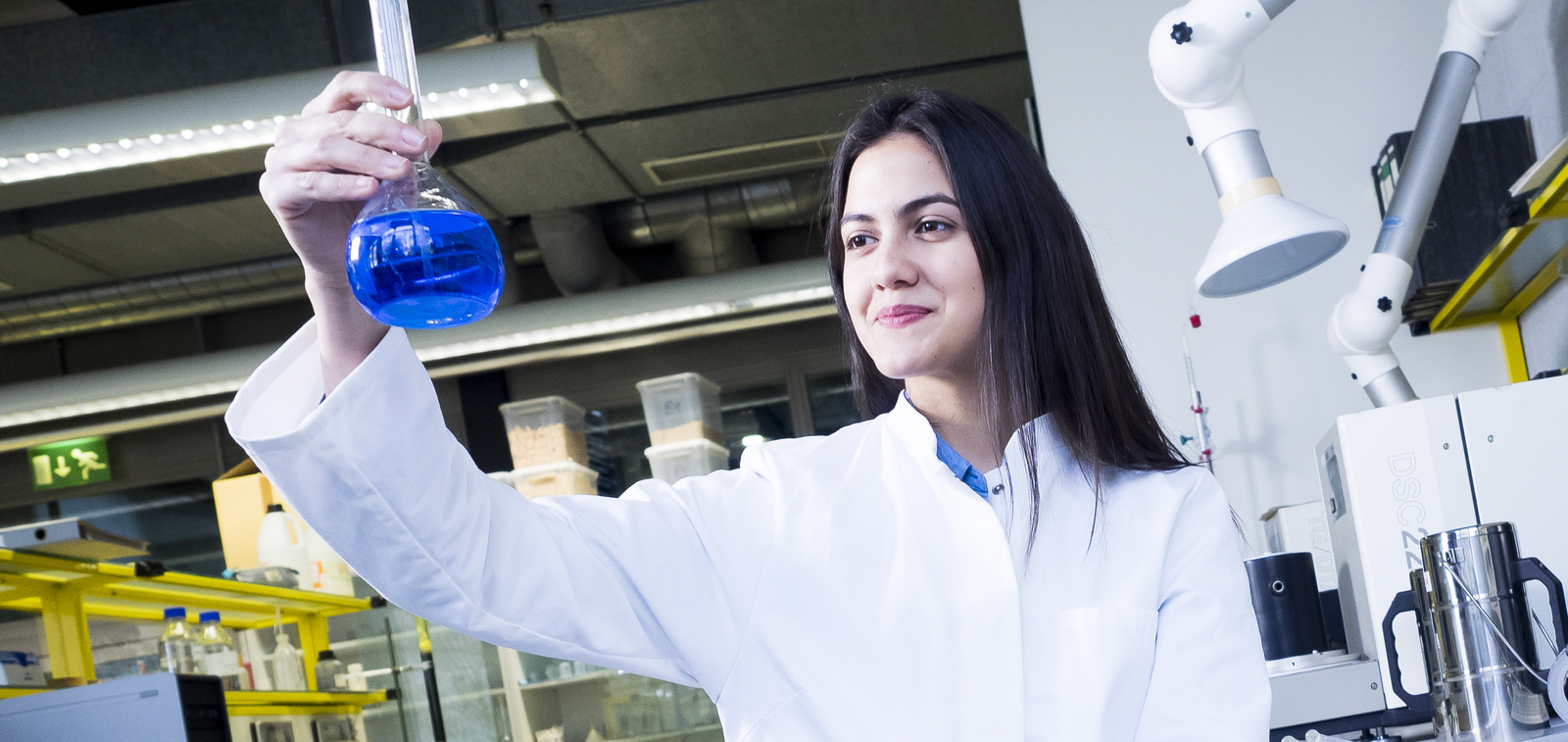 Frau im weißen Kittel schaut in einem Labor auf einen Reagenzkolben mit blauer Flüssigkeit