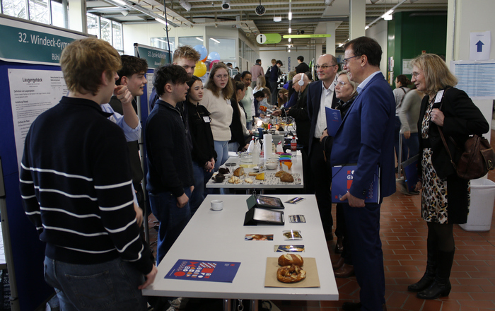 Mehrere Erwachsene stehen vor einem Tisch mit Laugengebäck und lassen sich von Schülerinnen und Schülern etwas erklären.