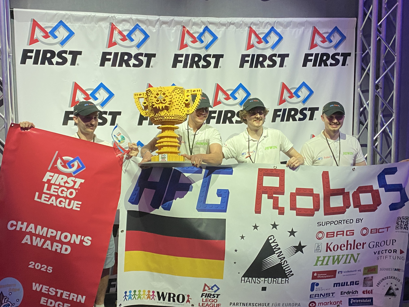 Vier junge Männer mit einem gelben Pokal aus Legosteinen und der Teamfahne