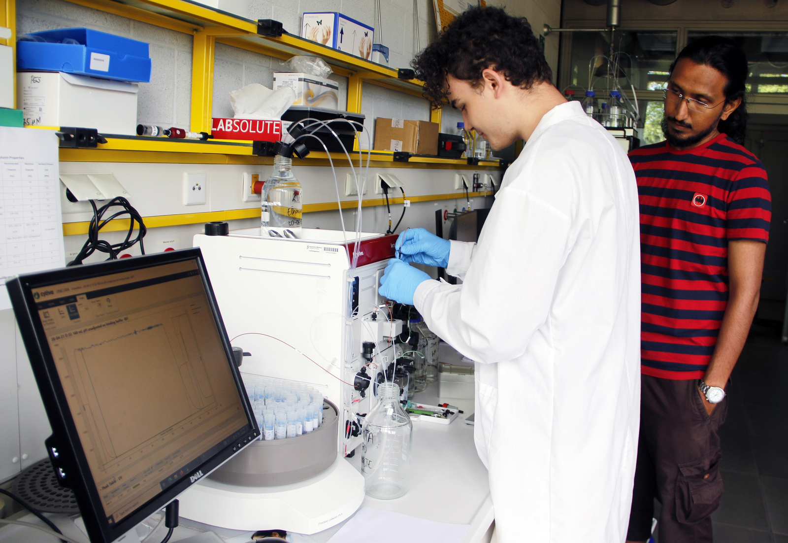 Gabriel Ben Freudenberg im weißen Kittel und mit blauen Handschuhen arbeitet beobachtet von Uzair Ahmed an einem Analysegerät